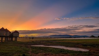 Die Silhouette von erhöhten Lodges vor einem atemberaubenden Sonnenaufgang über der weiten Savanne.