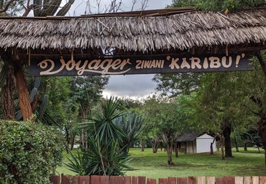 Ein hölzernes Schild mit der Aufschrift 'Voyager Ziwani Karibu!' begrüßt Besucher in einer grünen, bewaldeten Umgebung.