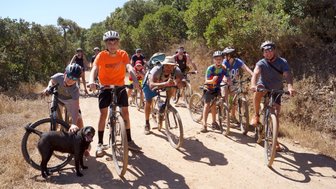 Eine Gruppe von Radfahrern, darunter Kinder und Erwachsene, posiert fröhlich auf einem staubigen Weg in der Natur.