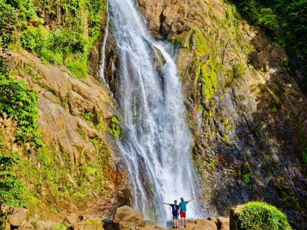 Zwei Personen stehen vor einem majestätischen Wasserfall, umgeben von üppigem, grünem Dschungel und Felsen.