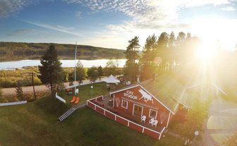 Ein rotes Ferienhaus mit einem weißen Pferdemotiv steht in einer malerischen Landschaft am Wasser, umgeben von Bäumen.