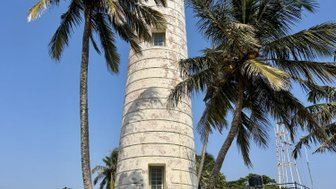Ein hoher Leuchtturm steht majestätisch zwischen Palmen unter einem strahlend blauen Himmel, umgeben von üppigem Grün.