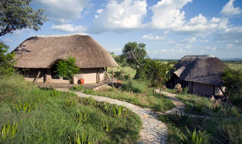 Zwei strohgedeckte Hütten stehen in einer grünen Landschaft, umgeben von Bäumen und sanften Hügeln unter einem blauen Himmel.