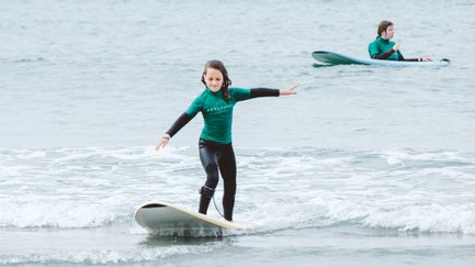 Ein Mädchen surft fröhlich auf einem Surfbrett, während ein anderer Surfer im Hintergrund im Wasser sitzt.
