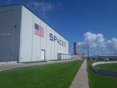Ein klarer Himmel über dem SpaceX-Gebäude, das mit der amerikanischen Flagge geschmückt ist, und einer Raketenstartanlage im Hintergrund.