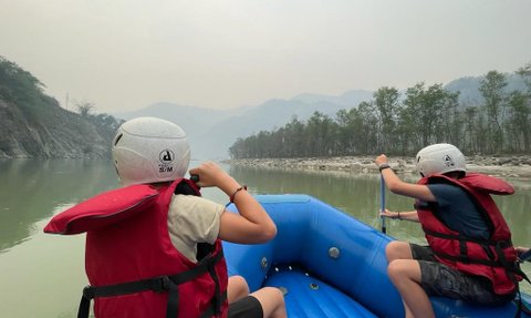 Zwei Kinder in roten Schwimmwesten paddeln in einem blauen Schlauchboot auf einem ruhigen Fluss in Nepal.
