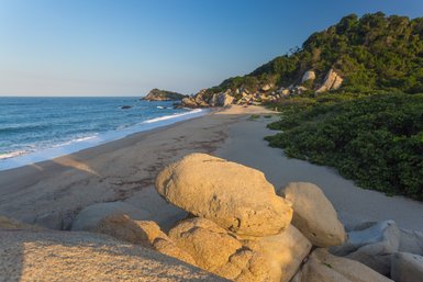 Ein ruhiger Strand mit sanften Wellen, umgeben von großen Felsen und üppigem Grün im Hintergrund, unter klarem Himmel.
