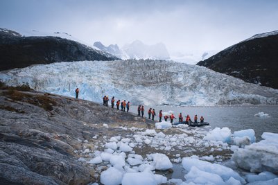Eine Gruppe von Touristen in orangefarbenen Schwimmwesten steht an einem glazialen Ufer, umgeben von Eisblöcken und Bergen.