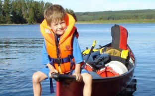Ein fröhlicher Junge sitzt auf der Kante eines Kanus, trägt eine orangefarbene Schwimmweste und genießt den sonnigen Tag am Wasser.
