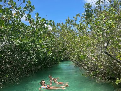 Drei Personen schwimmen entspannt in klarem Wasser, umgeben von üppigem Grün und strahlend blauem Himmel.