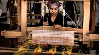 Eine Frau arbeitet konzentriert an einem Webstuhl und kreiert farbenfrohe Stoffe mit traditionellen Mustern.