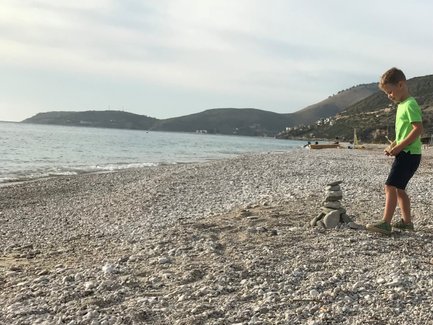 Ein Junge in einem grünen T-Shirt baut einen Steinturm am Kiesstrand, während das ruhige Wasser im Hintergrund glitzert.