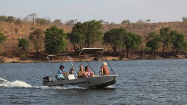 Eine Gruppe von fünf Personen genießt eine Bootsfahrt auf einem ruhigen Gewässer, umgeben von grünen Bäumen und trockenem Gelände.