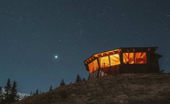 Eine beleuchtete Hütte steht auf einem Hügel, umgeben von dunklem Himmel und funkelnden Sternen.