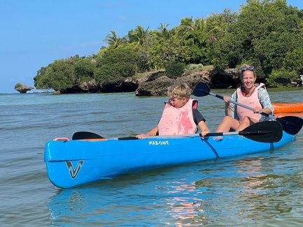 Ein Junge und eine Frau paddeln fröhlich in einem blauen Kajak, umgeben von klarem Wasser und üppigem Grün.