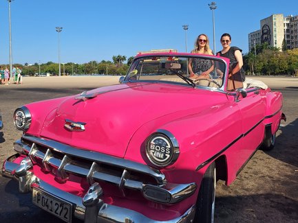 Zwei Frauen stehen lächelnd neben einem leuchtend pinken Oldtimer-Cabrio unter einem strahlend blauen Himmel.