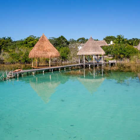 Ein ruhiger Wasserblick auf eine Holzplattform mit einem Strohdach, umgeben von klarem, türkisfarbenem Wasser und üppiger Vegetation.