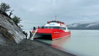 Ein rotes Boot liegt an einem sandigen Ufer, während Passagiere an Bord gehen und die ruhige Wasseroberfläche reflektiert.
