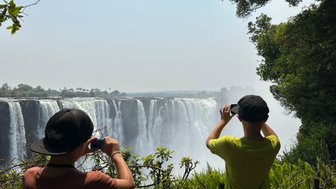 Zwei Kinder stehen am Rand eines Wasserfalls und fotografieren die beeindruckende Aussicht auf die Victoriafälle.