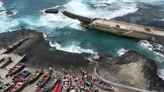 Bunte Fischerboote sind am Ufer eines lebhaften Wassers mit Wellen angedockt, während Menschen Aktivitäten im Hintergrund unternehmen.