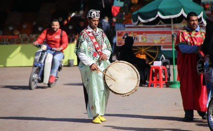 Ein Trommler in traditioneller Kleidung spielt auf einer Trommel, während Passanten in einem belebten Markt vorbeigehen.