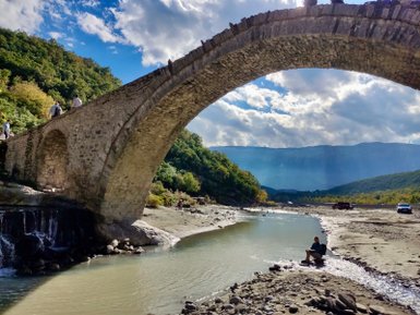 Ein alter Steinbogenbrücke überspannt einen ruhigen Fluss, während Menschen auf der Brücke und am Ufer spazieren.