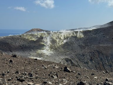 Ein rauchender Vulkan mit einer kargen, steinigen Landschaft und dem blauen Meer im Hintergrund unter einem klaren Himmel.