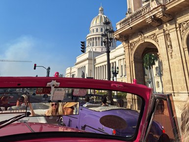 Im Vordergrund sind fröhliche Passagiere in einem pinken Cabrio zu sehen, während sie an historischen Gebäuden von Havanna vorbeifahren.