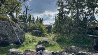 Eine malerische Landschaft mit einem Lagerfeuerplatz, umgeben von großen Steinen und üppigem Grün unter einem bewölkten Himmel.