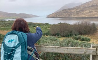 Eine Person mit einem Rucksack zeigt auf die ruhige Killary Fjord-Landschaft, umgeben von sanften Hügeln und bewaldeten Ufern.