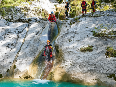 Ein Kind rutscht fröhlich einen glatten Felsen hinunter, während andere Canyoning-Teilnehmer zuschauen und anfeuern.
