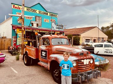 Ein Junge steht vor einem alten, rostigen Lastwagen vor einem bunten Geschäft in Seligman, Arizona, mit Wolken am Himmel.