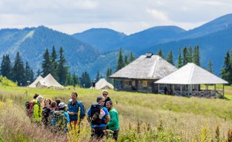 Eine Gruppe von Wanderern steht in einer blühenden Wiese, umgeben von Bergen und Zelten, und diskutiert lebhaft.