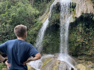 Ein Junge steht am Ufer eines klaren Wassers, während ein beeindruckender Wasserfall hinter ihm herabstürzt.