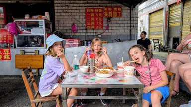 Drei Mädchen sitzen an einem Tisch und genießen asiatisches Essen, während sie mit Stäbchen essen und fröhlich wirken.
