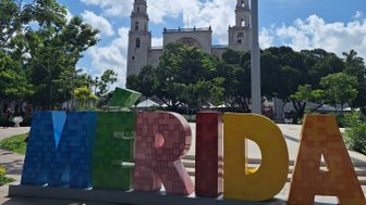 Bunte Buchstaben bilden das Wort 'Mérida' vor einer beeindruckenden historischen Architektur unter einem strahlend blauen Himmel.