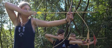 Eine Gruppe von Frauen zielt mit Pfeil und Bogen im Freien, umgeben von dichter Natur und Lichtstrahlen zwischen den Bäumen.