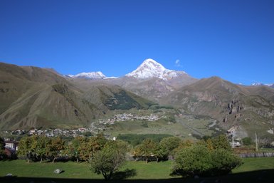 Im Vordergrund stehen grüne Wiesen mit Bäumen, während majestätische Berge im Hintergrund mit Schnee bedeckt sind.