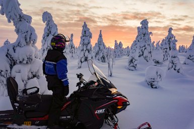 Ein Schneemobilfahrer steht in einer verschneiten Landschaft, umgeben von schneebedeckten Bäumen und einem farbenfrohen Sonnenuntergang.