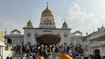 Eine Gruppe von Menschen mit orangefarbenen Turbanen versammelt sich vor dem Gurudwara Bangla Sahib, das prächtig im Hintergrund steht.