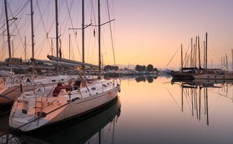 Eine ruhige Marina bei Sonnenaufgang, mit mehreren Segelbooten, die sanft im Wasser schaukeln und ihre Spiegelungen zeigen.