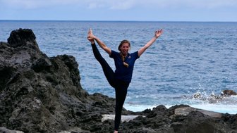 Eine fröhliche Frau macht eine Yoga-Pose auf einem Felsen am Meer, mit einem strahlenden Lächeln und erhobenen Armen.