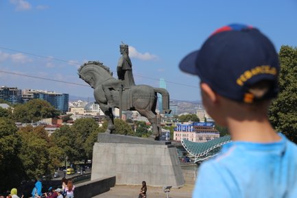 Ein Junge mit einer blauen Mütze beobachtet eine beeindruckende Statue eines Reiters in Tiflis, umgeben von Stadtlandschaft.