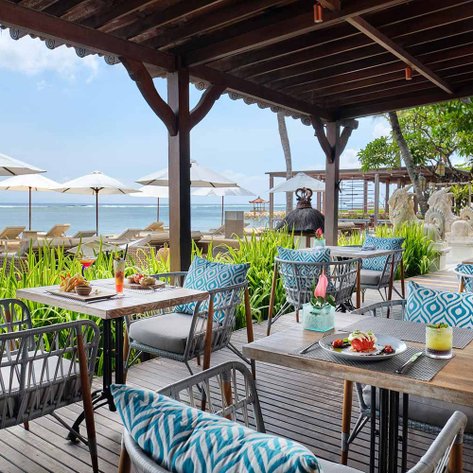 Ein einladendes Restaurant am Strand mit stilvollen Tischen und Stühlen, umgeben von Palmen und Sonnenschirmen.