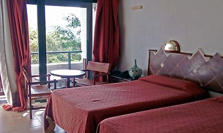 Ein helles Zimmer mit zwei Einzelbetten, roten Bettüberzügen und einem kleinen Tisch mit Stühlen neben einem großen Fenster.