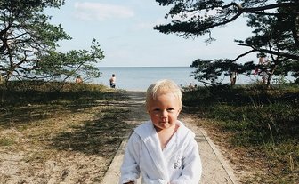 Ein kleines Kind in einem weißen Bademantel läuft barfuß auf einem Weg zum Strand, umgeben von Bäumen und Wasser.