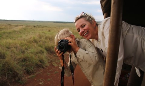 Ein fröhlicher Junge mit einer Kamera posiert, während eine lächelnde Frau ihm zur Seite steht, umgeben von einer weiten Savanne.