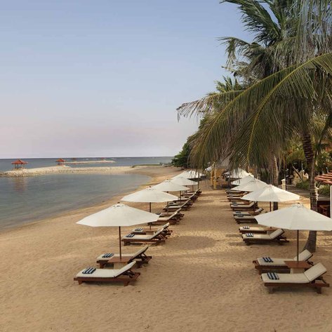 Ein ruhiger Strand mit Liegen und Sonnenschirmen, umgeben von Palmen und einem klaren blauen Himmel über dem Wasser.
