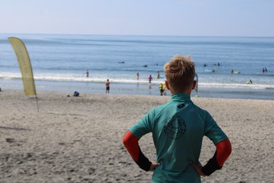 Ein Junge in einem grünen Surfanzug beobachtet das Meer, während andere im Wasser surfen und spielen.