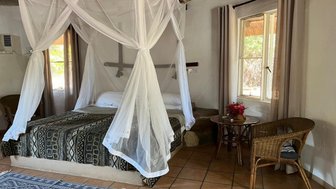 Ein gemütliches Schlafzimmer mit einem großen Bett, umgeben von einem Moskitonetz und einem rustikalen, geflochtenen Stuhl.
